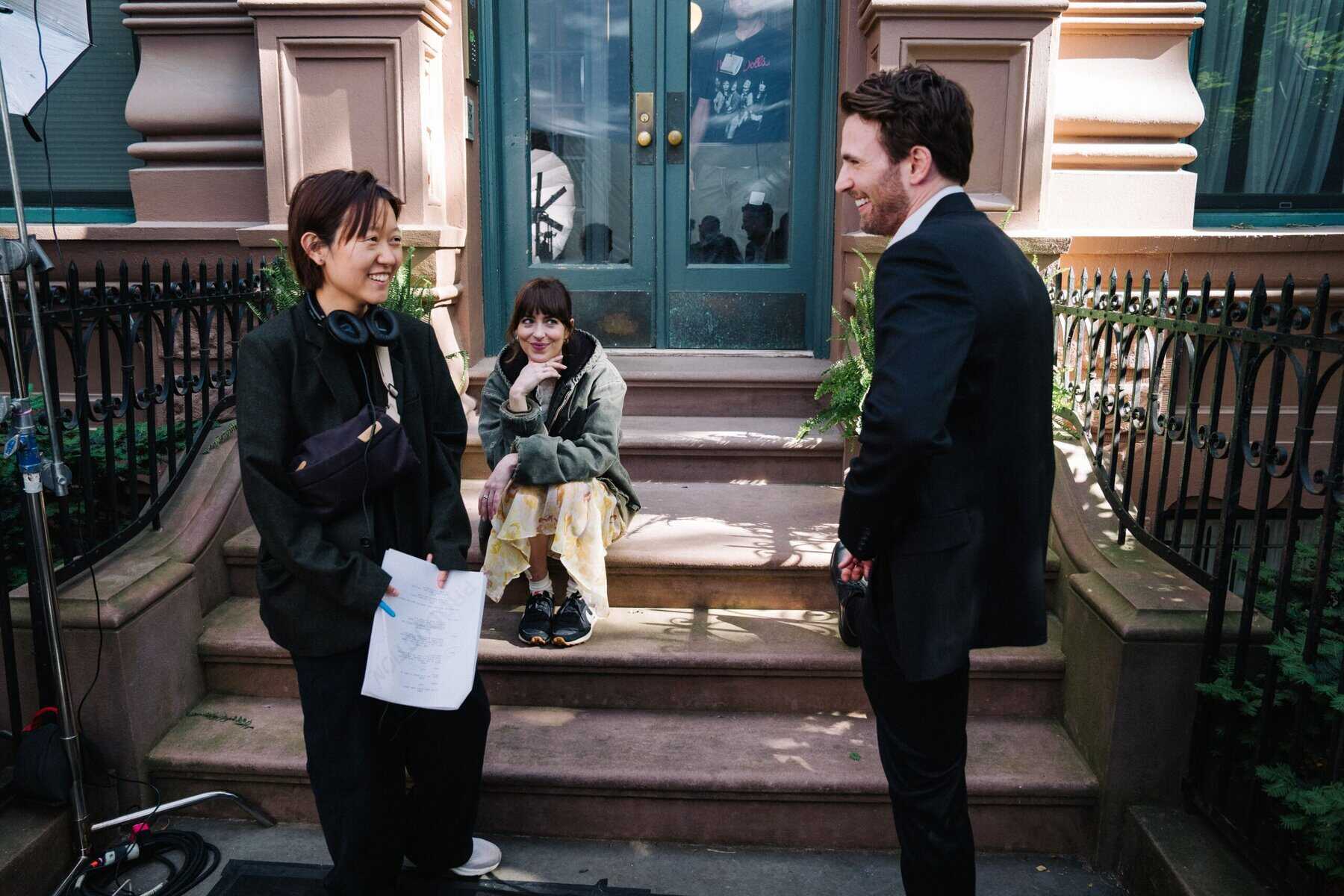 Friendly young woman holding script, professional man in suit, and excited woman sitting on steps outside building, promoting film or media project. L–R) Director Celine Song with Dakota Johnson and Chris Evans on the set of Materialists (2024). Photo by Atsushi Nishijima. © A24. Image courtesy of A24 Press.